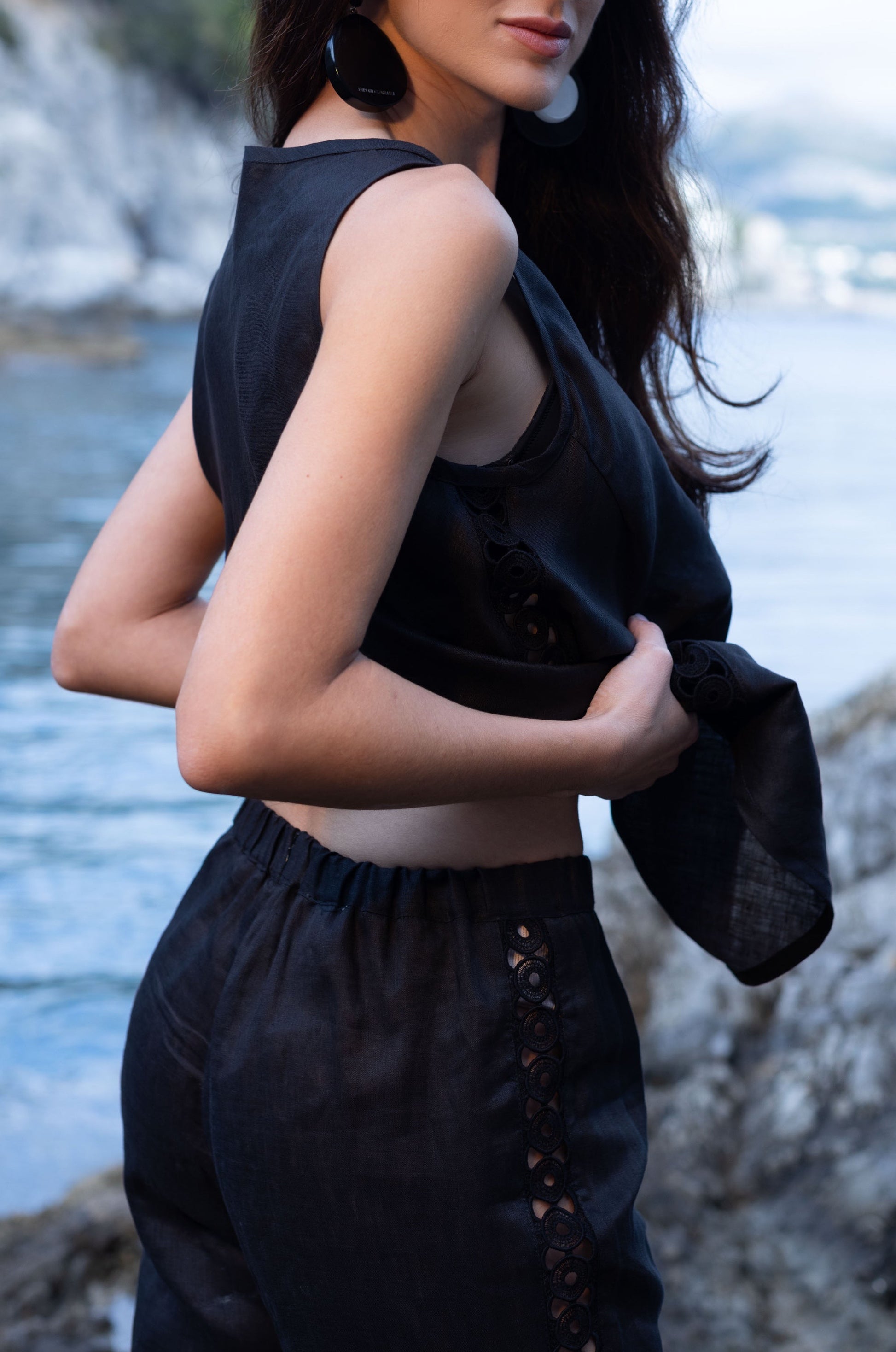 Woman in Black sleeveless top and pants standing by a body of water with rocky cliffs in the background.