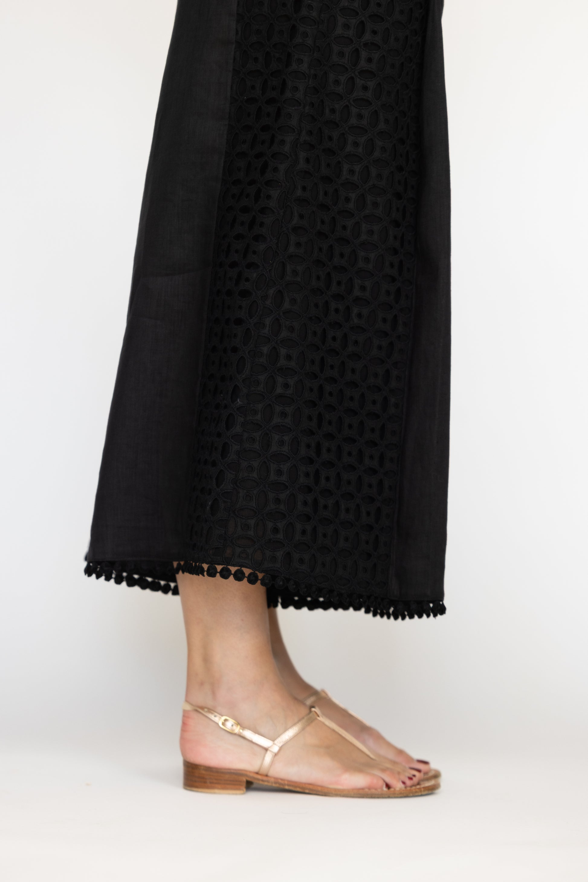 Black pants with a geometric pattern and tassel trim worn by a person, paired with gold sandals on a background.