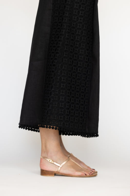 Black pants with a geometric pattern and tassel trim worn by a person, paired with  sandals on a background.